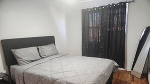 Een net opgemaakt bed met een grijze deken, een donker hoofdbord en een zwart gordijn met patroon.