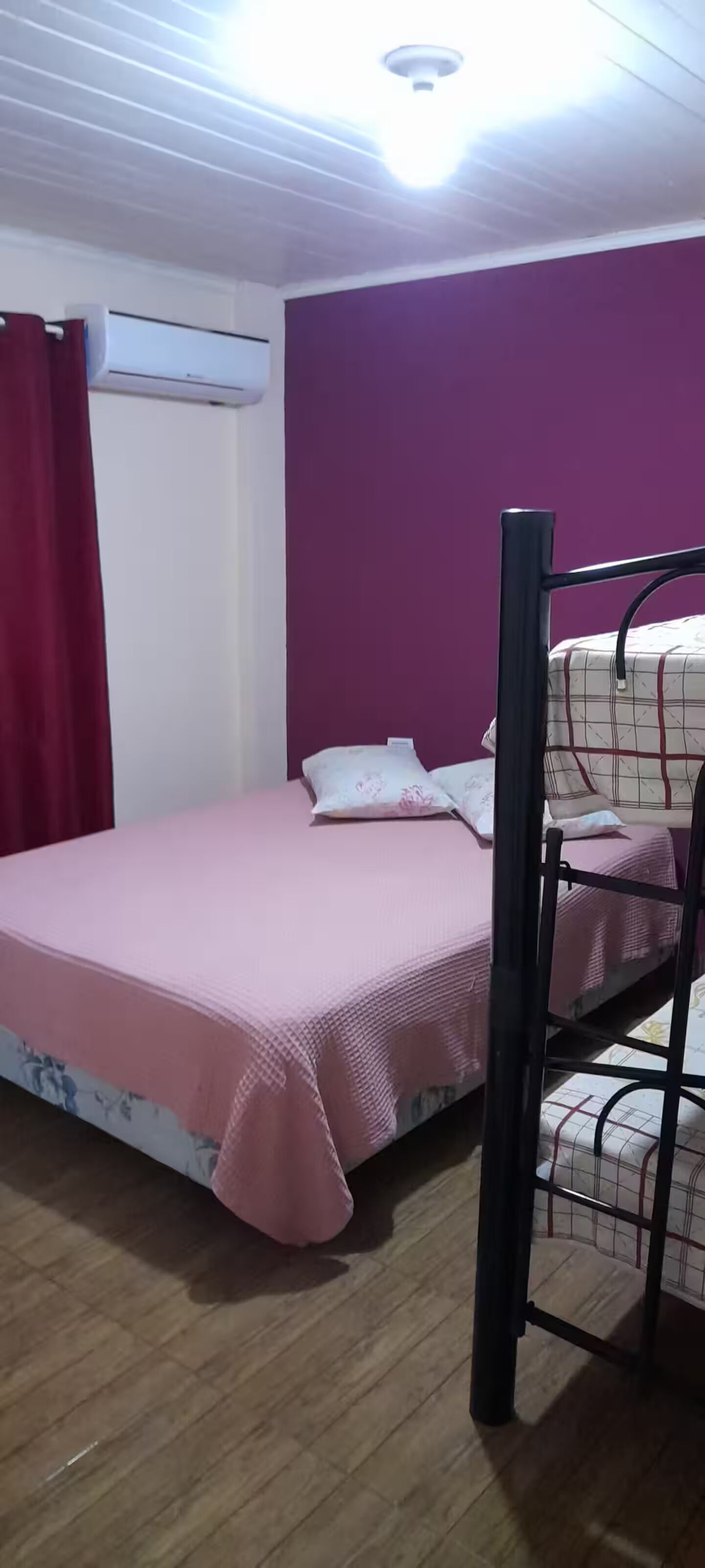Une chambre avec un lit superposé, un lit simple recouvert d’une couverture rose, un rideau rouge et un climatiseur fixé au mur.