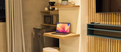 A compact, modern bathroom with a built-in shower, a small desk with a laptop, a microwave, and a shelf with decorative items.