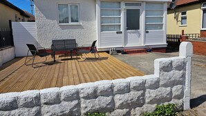 Une petite maison avec une terrasse en bois, deux chaises noires et une porte blanche.