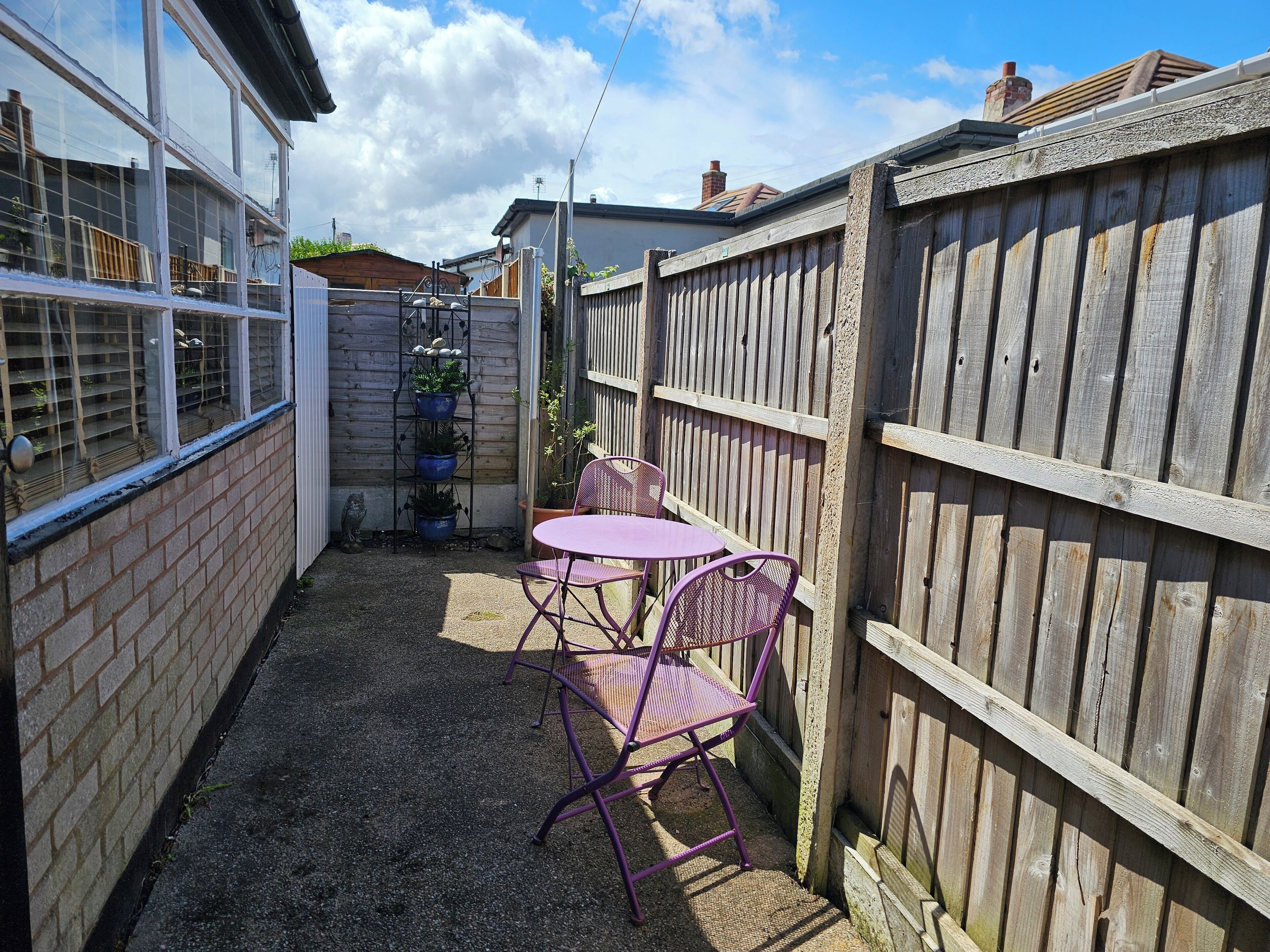 A narrow outdoor patio with a brick wall, a wooden fence, and a metal chair and table.