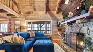 A cozy log cabin interior with a large blue sectional sofa, a stone fireplace, and a wooden staircase. - Cozy 4-bedroom cabin with separate living spaces, and all the amenities. (Sevierville)