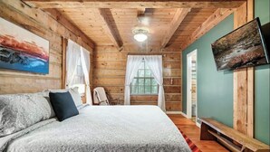 A wooden cabin bedroom with a bed, a bench, a TV, and a window with curtains. - Cozy 4-bedroom cabin with separate living spaces, and all the amenities. (Sevierville)