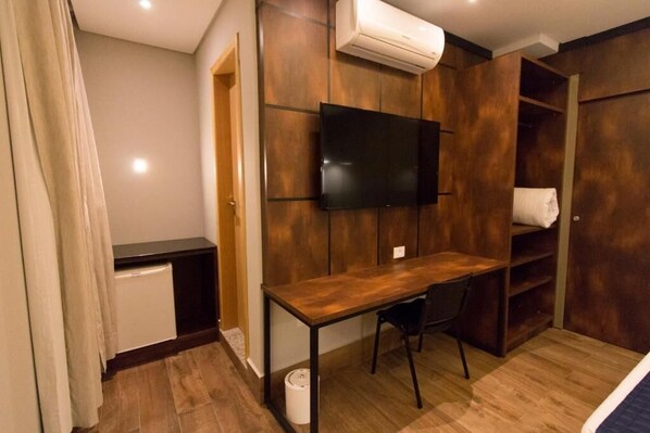 A compact room with a wooden desk, a chair, a television mounted on the wall, and a wardrobe with a towel hanging inside. - Adria Premium Hotel (Guarapuava)