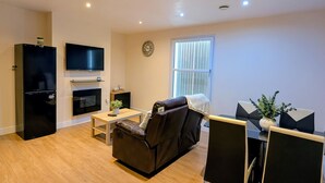 A modern living room with a black leather sofa, a dining table with chairs, a black refrigerator, a flat-screen TV mounted on the wall, and a window with blinds. - Upperhead Row Apartment (Huddersfield)