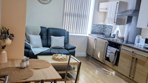 A modern living room with a black leather sofa, a wooden dining table with chairs, and a kitchen area with cabinets and a stove. - Upperhead Row Apartment (Huddersfield)