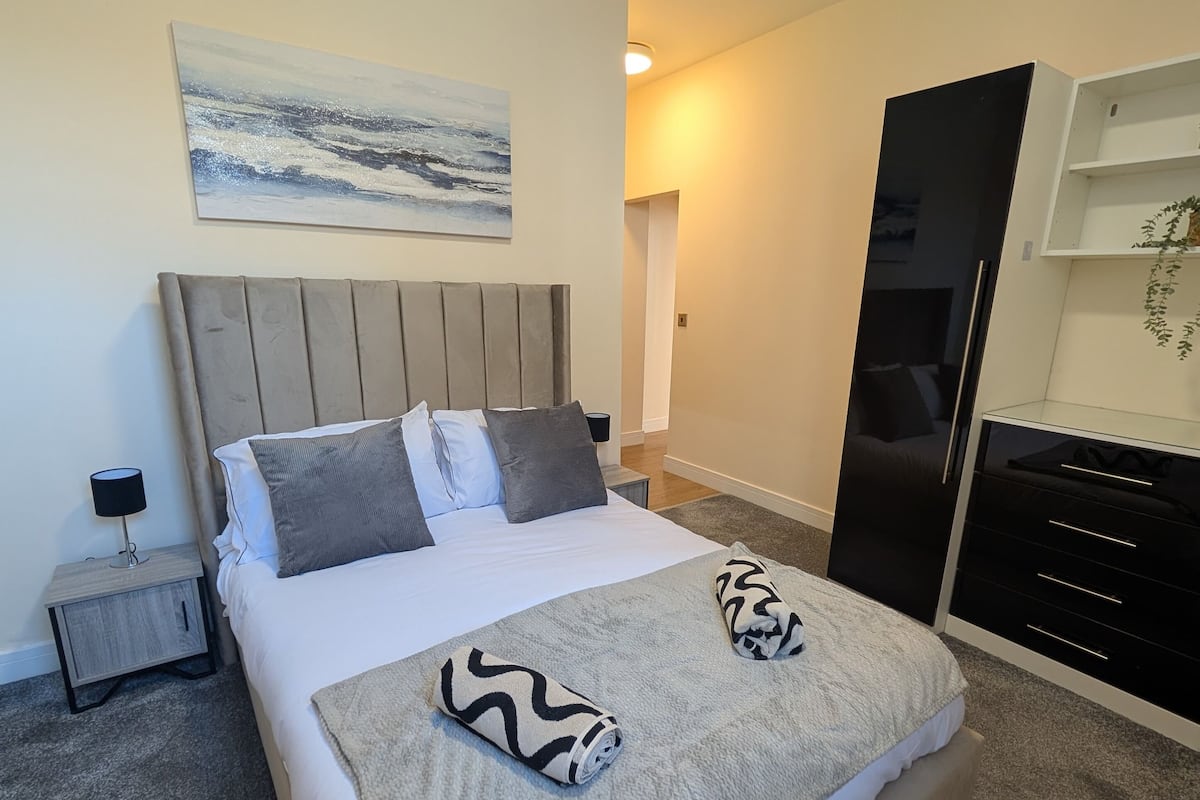 A neatly made bed with a headboard, a bedside table with a lamp, a wall-mounted shelf, and a painting.