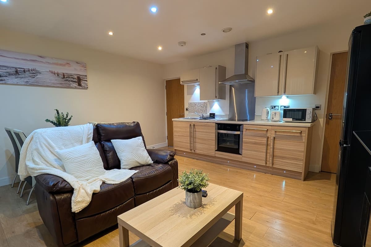 A modern living room with a brown leather sofa, a wooden coffee table, and a kitchen area with white cabinets and stainless steel appliances.