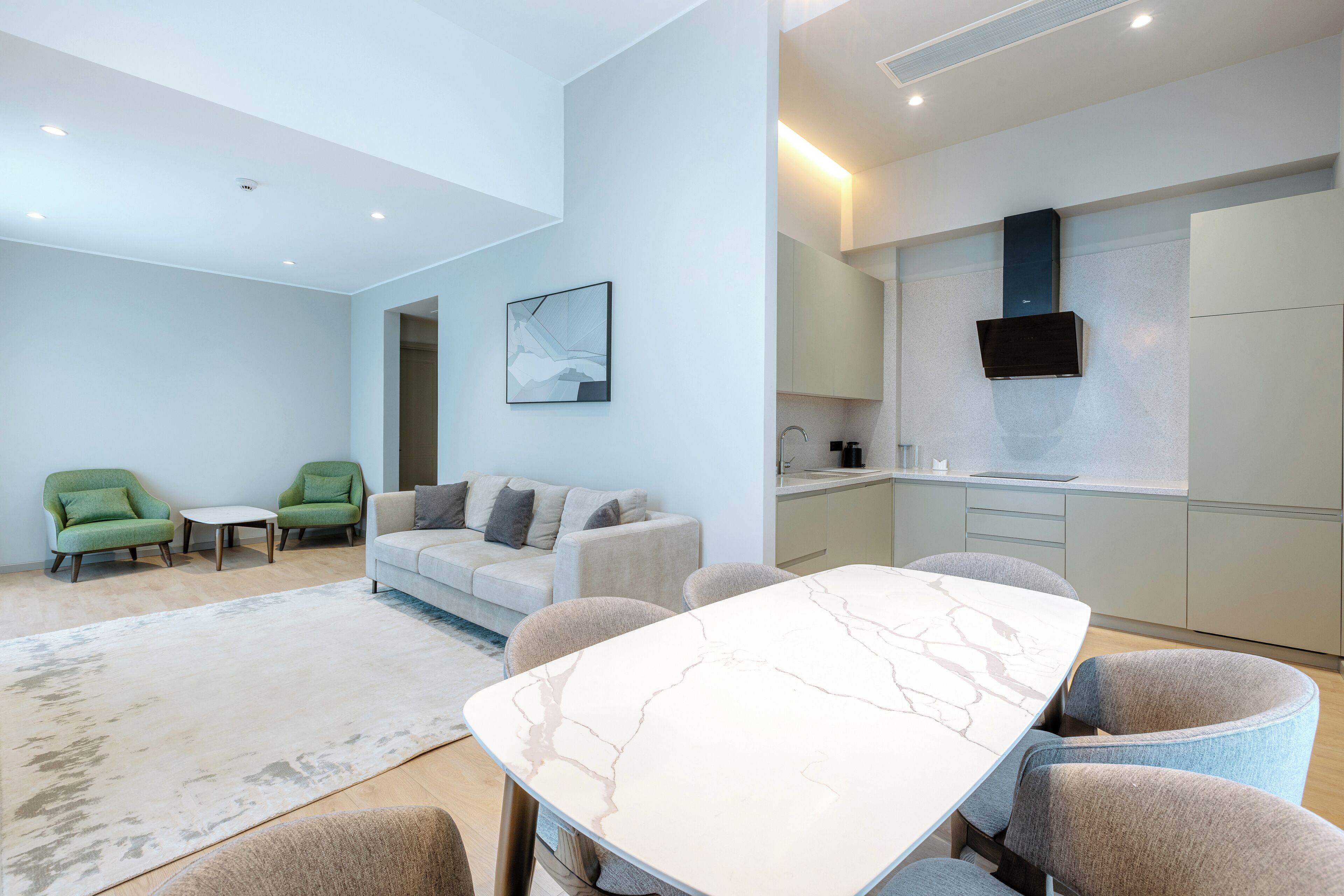 A modern living room with a marble dining table, green armchairs, a grey sofa, and a kitchen area with a built-in oven and a flat-screen TV.