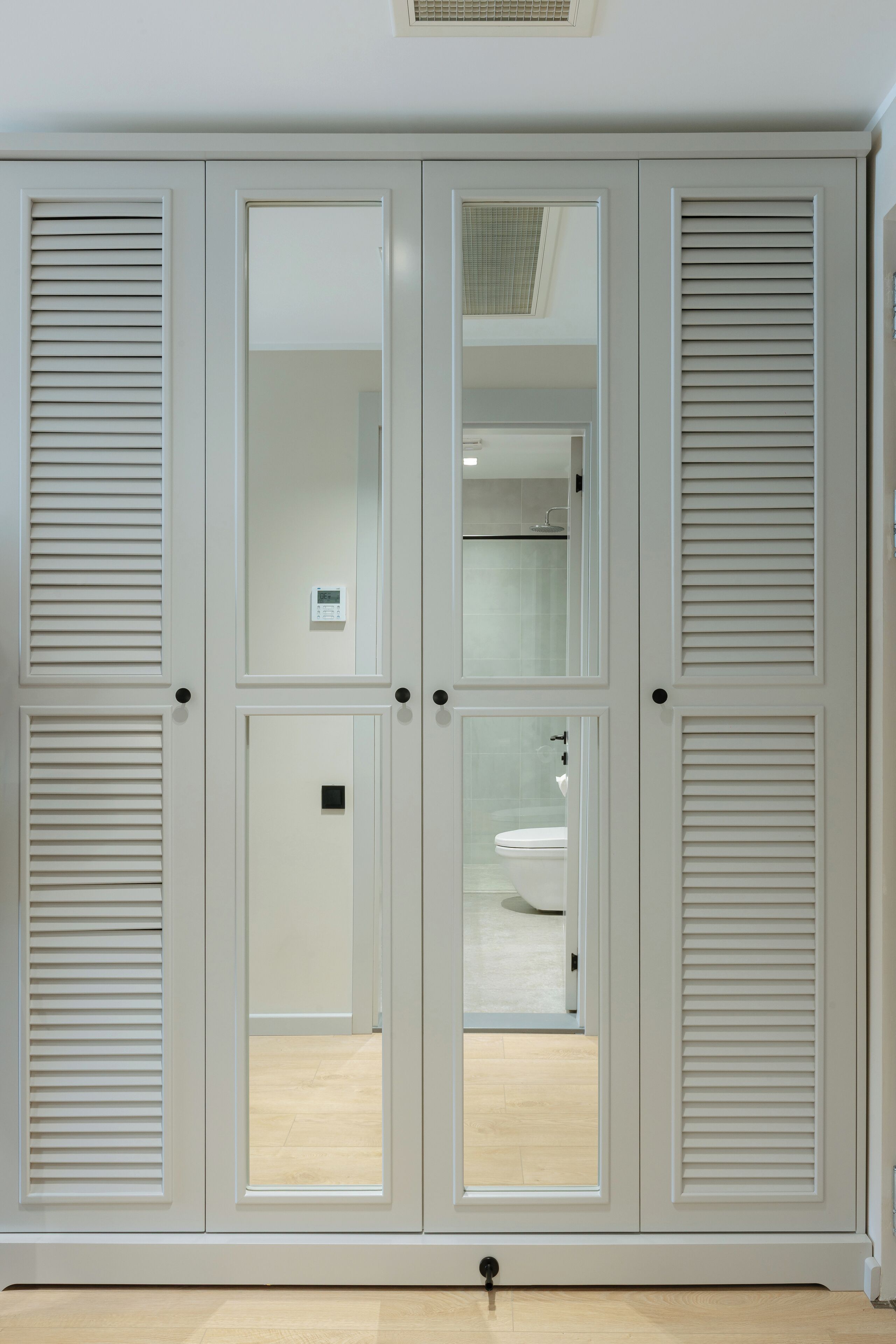 Un conjunto de puertas dobles blancas con paneles persiana, un baño visible con inodoro y lavamanos, y un piso de madera clara.