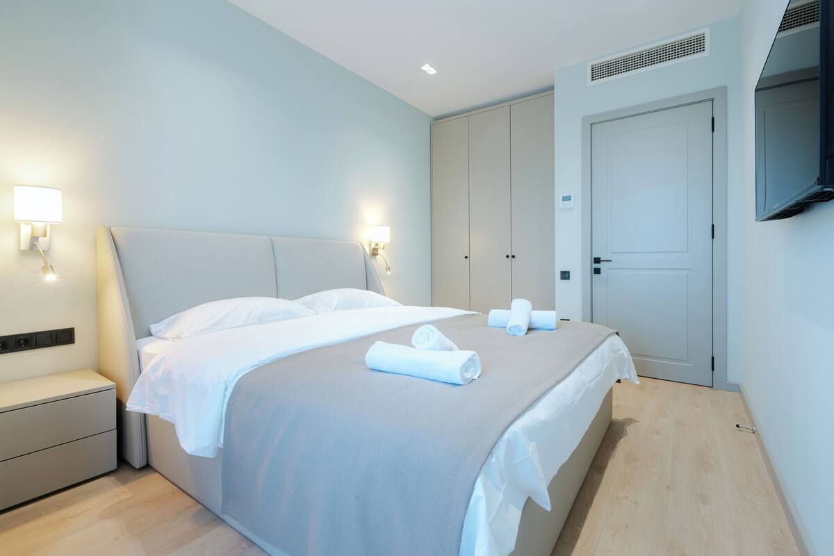 Ein modernes Hotelzimmer mit einem ordentlich bezogenen Bett, einem Nachttisch, einem Kleiderschrank und einem an der Wand befestigten Fernseher.