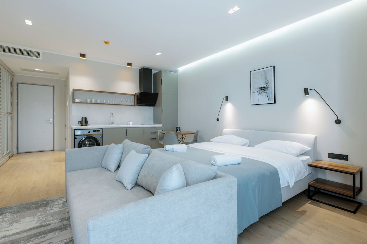 Ein modernes Hotelzimmer mit grauem Sofa, einem Bett mit weißen Leinentüchern, einem kleinen Holznachttisch und einer wandmontierten Lampe.
