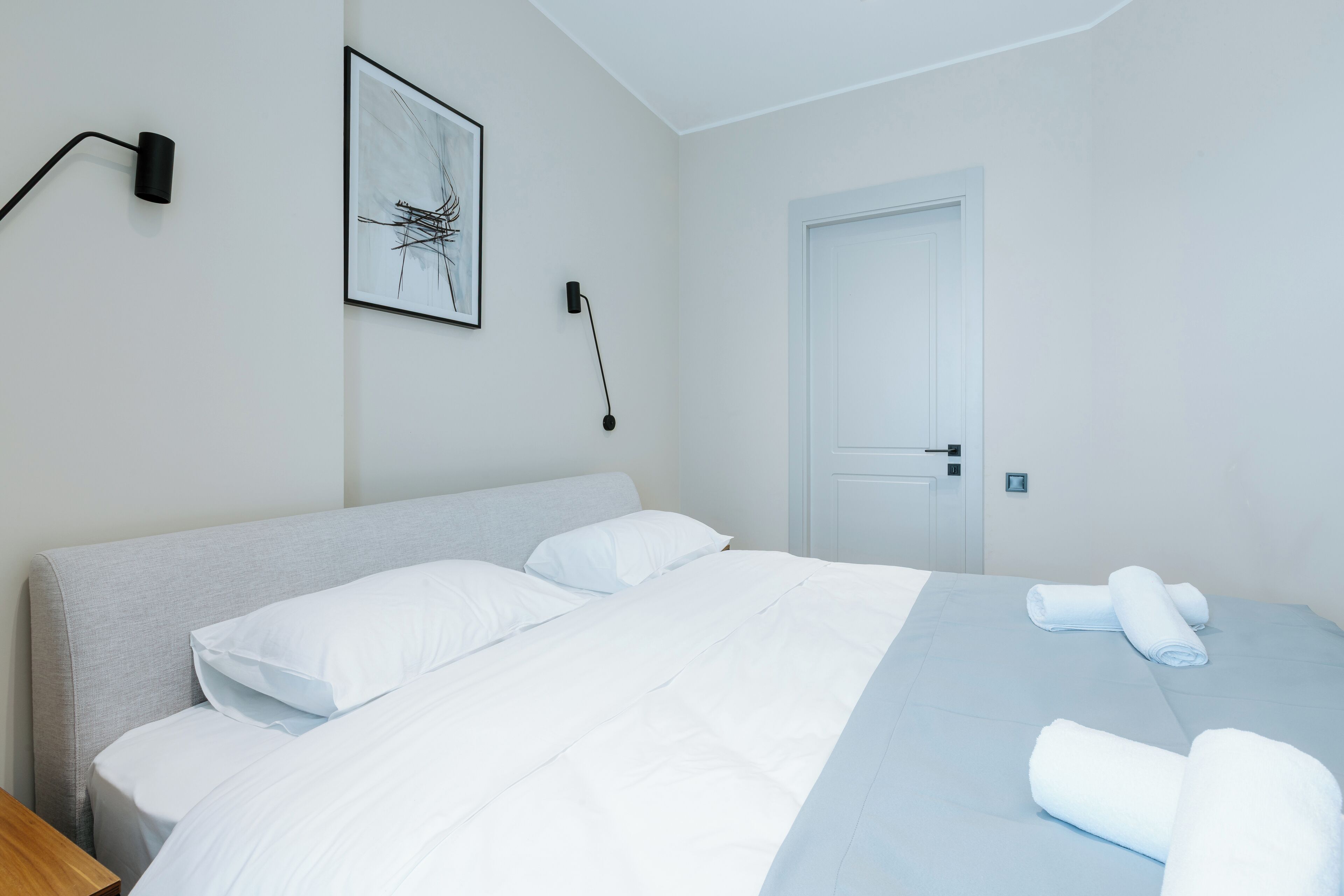 Un letto rifatto con lenzuola bianche e azzurre, una testiera grigia, un'opera d'arte astratta incorniciata al muro e una porta con maniglia.
