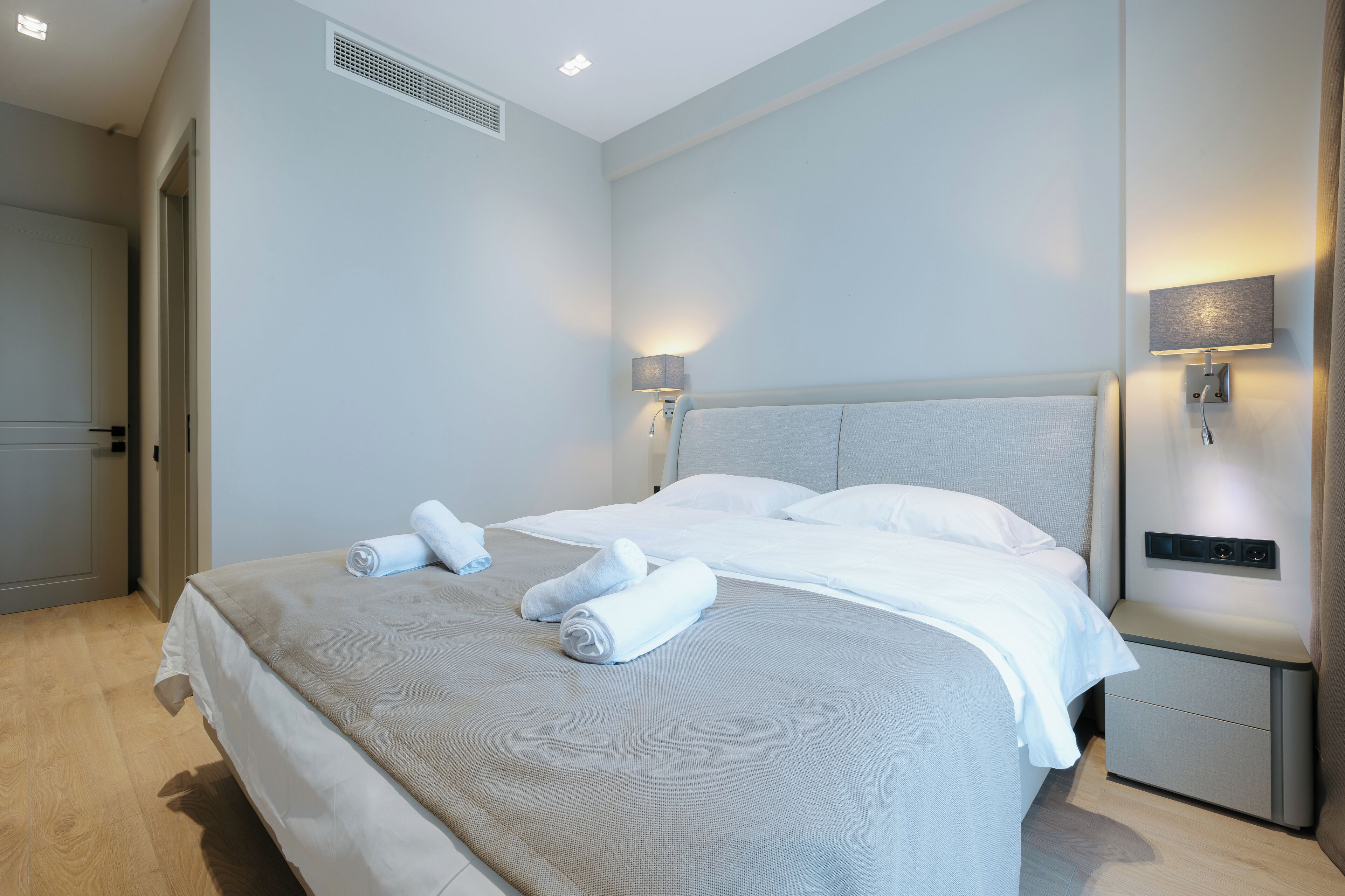 A neatly made bed with white linens and rolled towels, a bedside table with a lamp, and a wall-mounted light fixture.