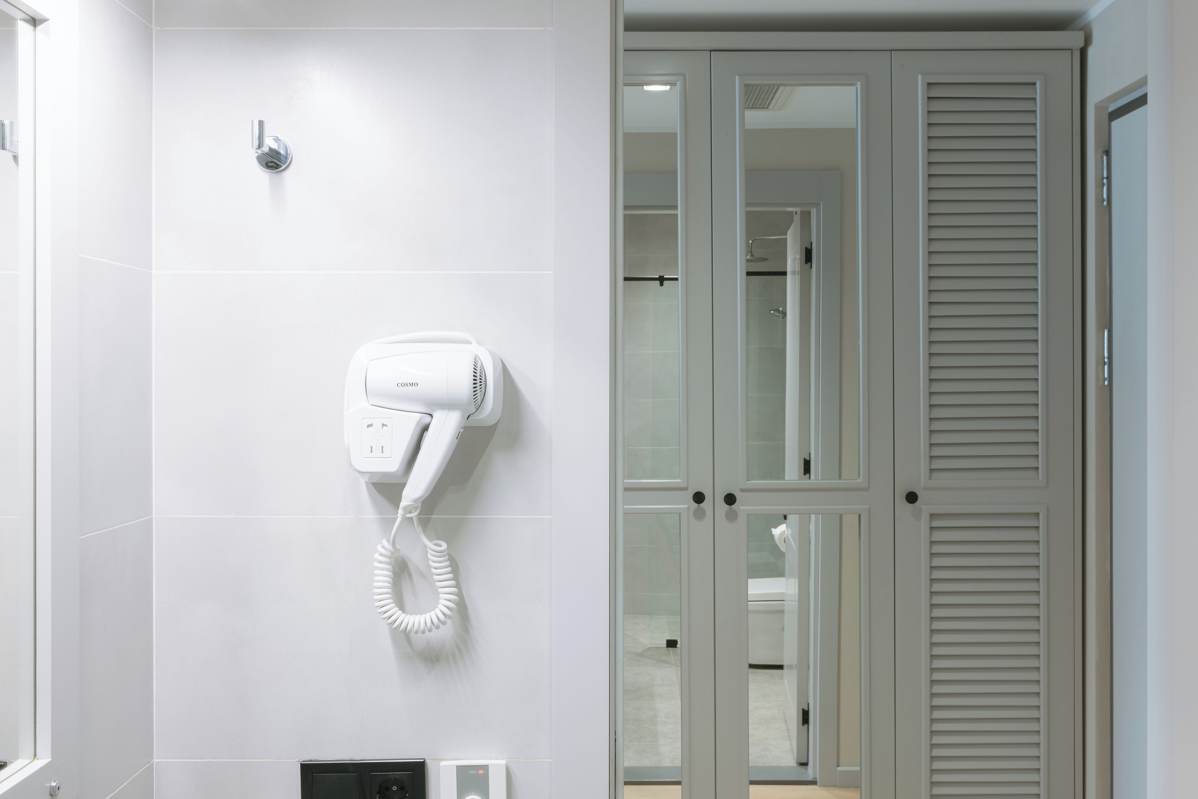 A modern bathroom with a hairdryer mounted on the wall, a glass shower enclosure, and a door with louvered panels.