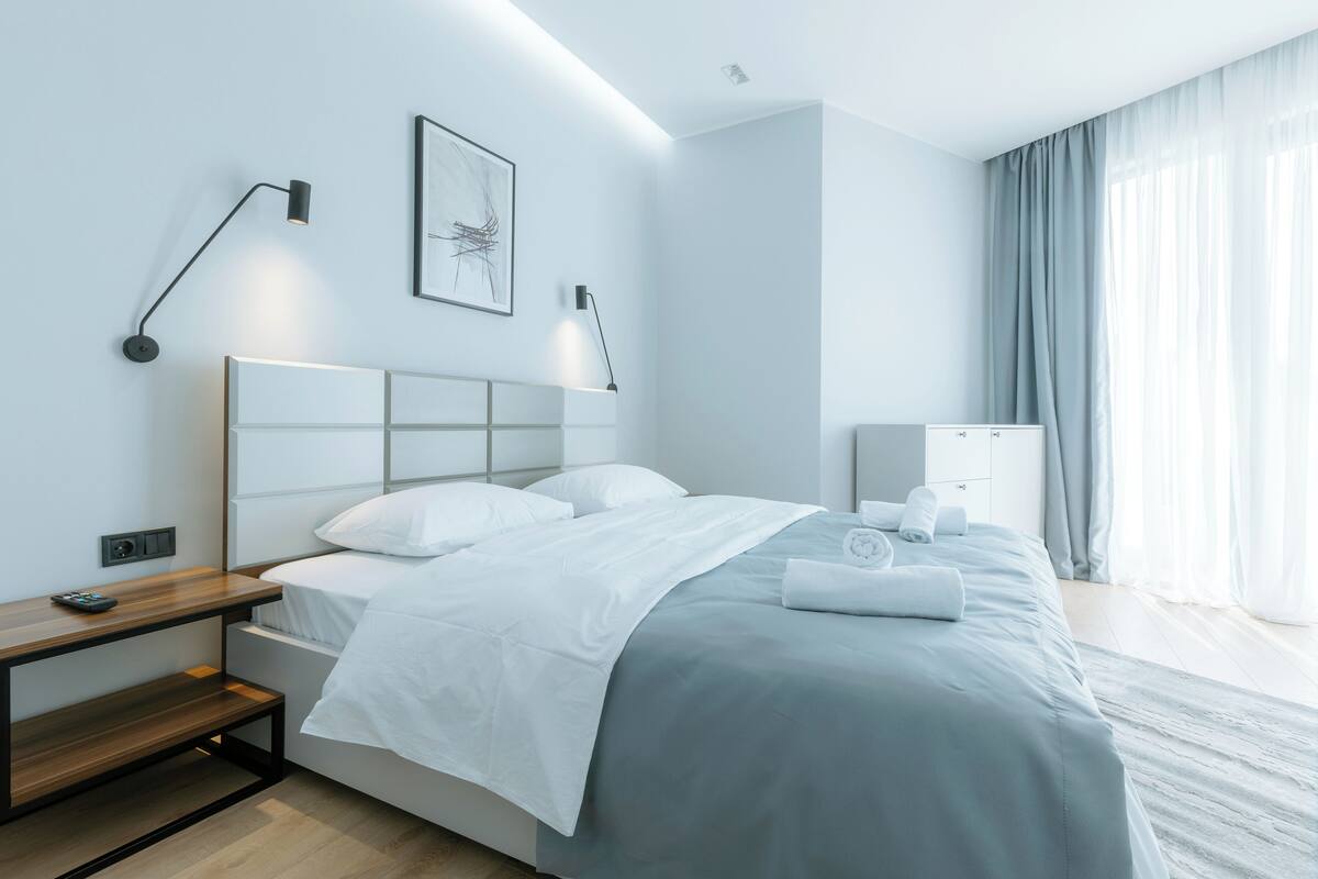 Ein modernes Hotelzimmer mit einem ordentlich bezogenen Bett, Nachttischen, einem Schreibtisch und einem großen Fenster mit Vorhängen.