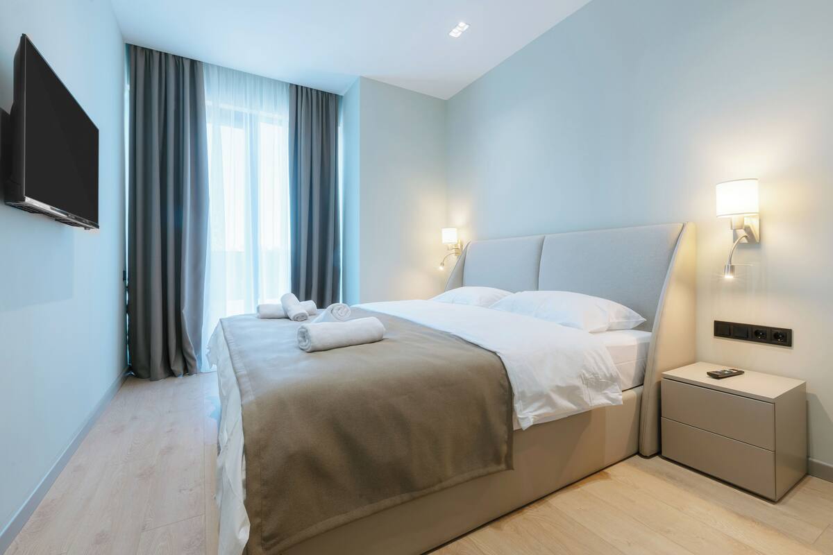 Ein modernes Hotelzimmer mit einem großen Bett, einem flachbildfernseher an der Wand, einem Nachttisch mit Lampe und einem fenster mit Vorhängen.