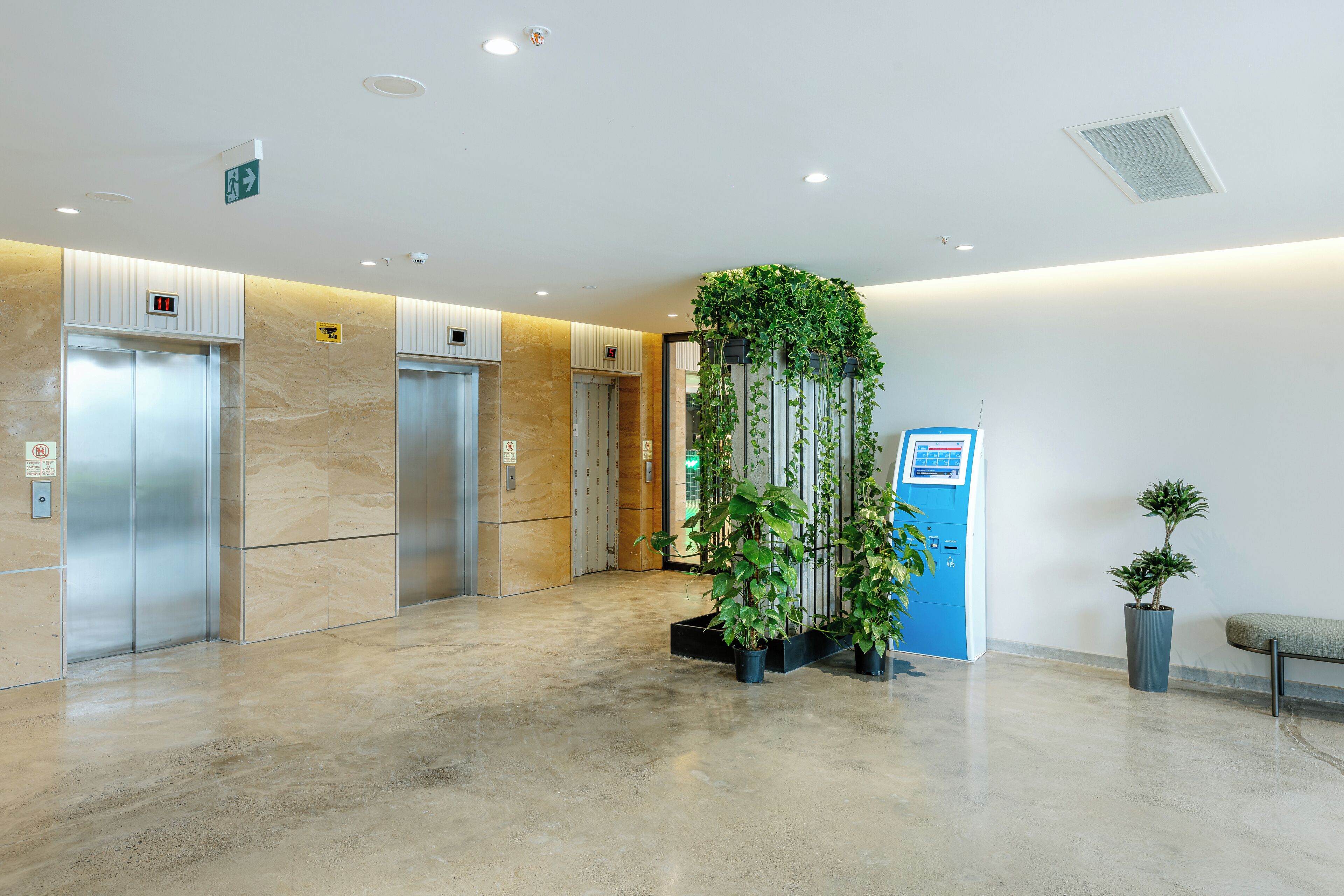 Un área de ascensores moderna con tres elevadores, un quiosco de información azul y plantas en macetas.