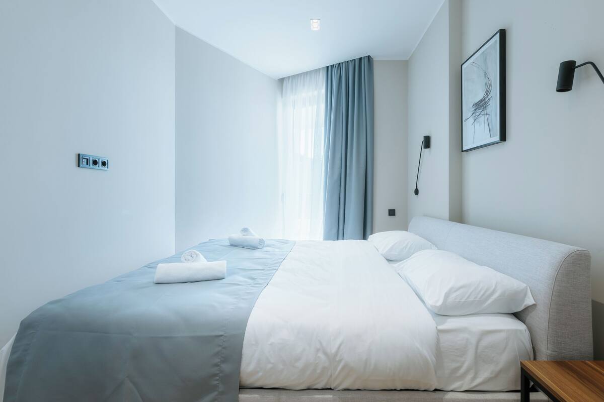 Ein modernes Hotelzimmer mit einem ordentlich bezogenen Bett, einem Nachttisch, einer Wandlampe und einem Fenster mit Vorhängen.