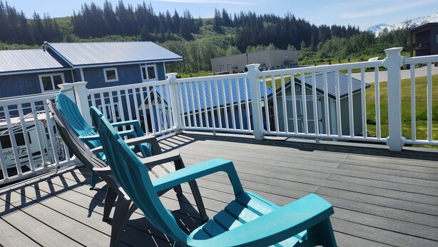 A deck with turquoise chairs and a white railing, overlooking a forested area with buildings in the background.