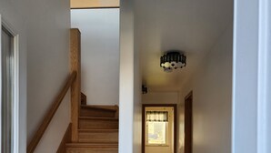 A hallway with a wooden staircase leading to a second floor. The hallway has a tiled floor and a ceiling light fixture.