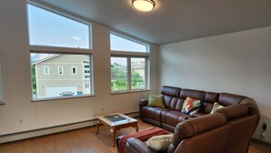 A spacious living room with a brown leather sofa, a wooden coffee table, and a view of a residential area through large windows.