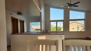 A spacious kitchen with large windows, a ceiling fan, and wooden chairs at a dining table.