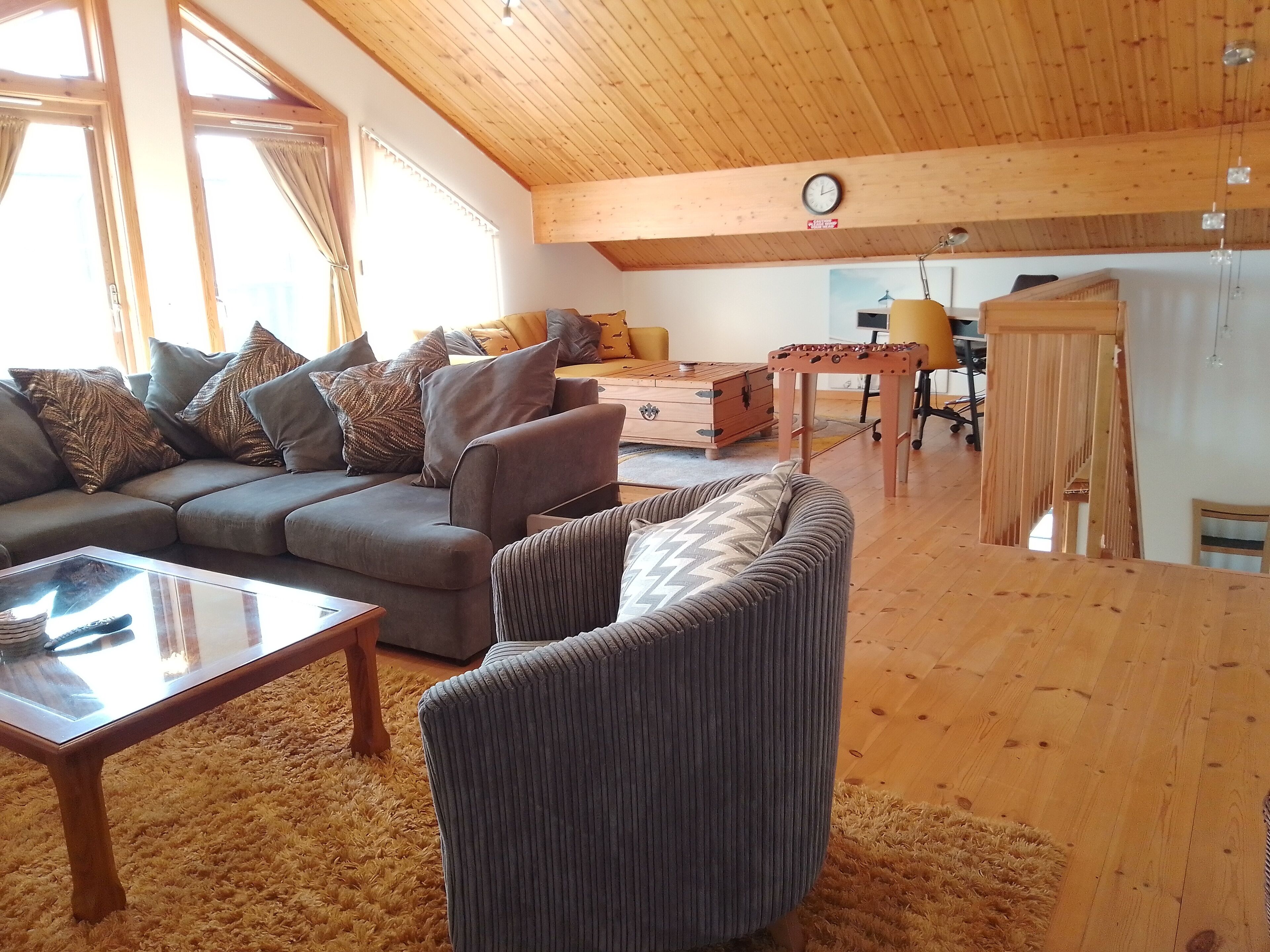 Uma sala de estar aconchegante com um sofá, poltrona e mesa de centro. Teto de madeira e um relógio na parede.
