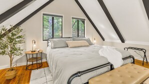 Un dormitorio con techo inclinado, una cama con colcha gris, dos ventanas, una mesita de noche con lámpara, una planta en maceta y un banco.