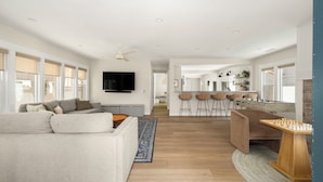 A spacious living room with a grey sofa, a wooden coffee table, and a blue rug. There is a kitchen area with bar stools and a foosball table. - Belmont Beach Escape: Private Pool and Hot Tub + Steps to the Sand (Long Beach)