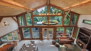 A spacious loft-style room with a high ceiling, large windows, a central chandelier, a fireplace, and a cozy seating area. - Private Tree Cabin on 8 Acres - King Suite, Large Deck & Fire Pit (Tomball)