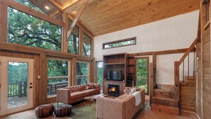 A spacious living room with a large fireplace, wooden beams, and a view of the outdoors. - Private Tree Cabin on 8 Acres - King Suite, Large Deck & Fire Pit (Tomball)