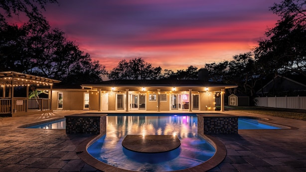 A pool area with a circular hot tub, a covered patio, and a house with multiple windows and lights.