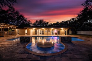 A pool area with a circular hot tub, a covered patio, and a house with multiple windows and lights. - Indian Rocks Escape (Largo)