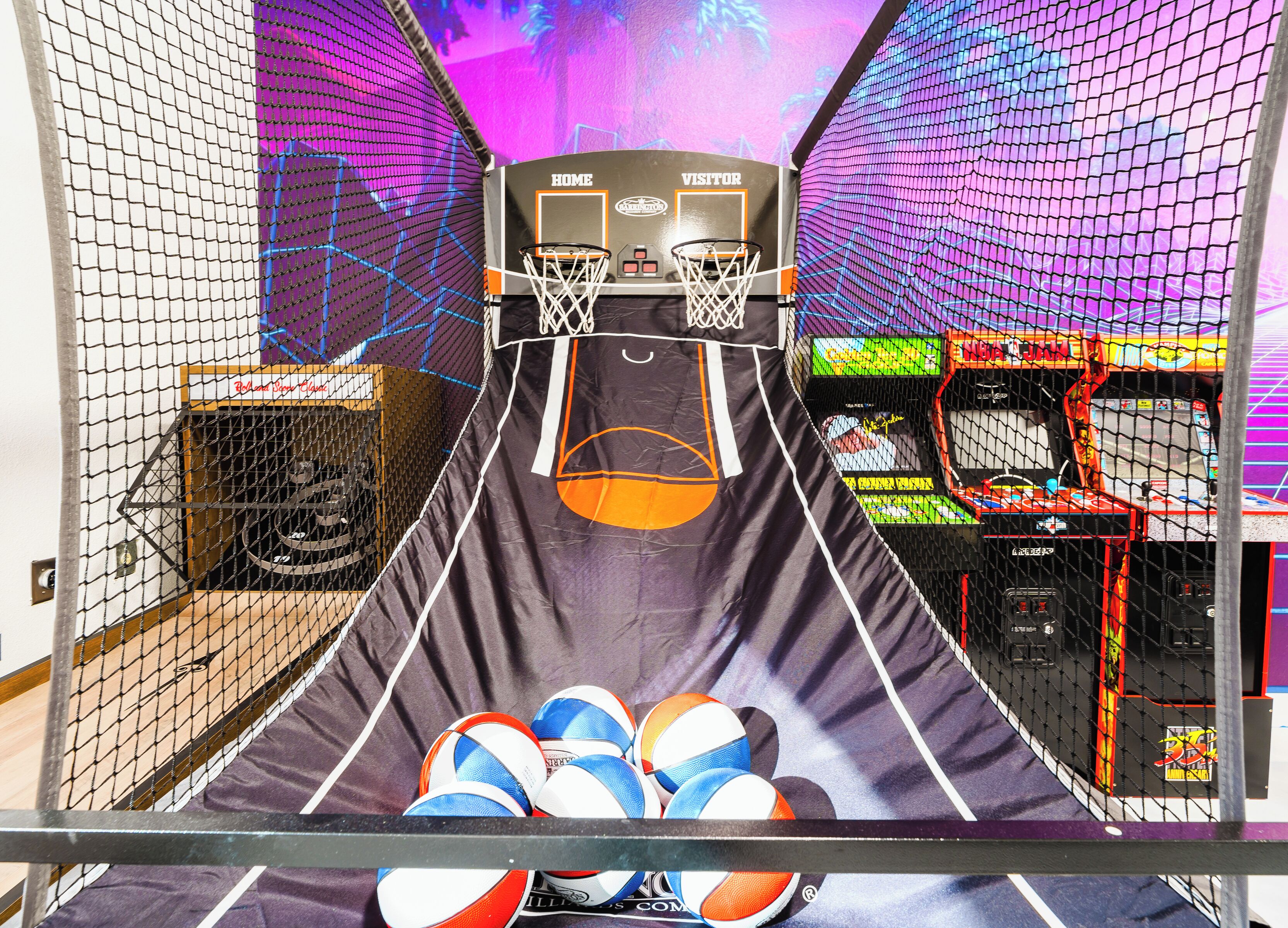Um salão de jogos de basquete com área de jogo cercada por rede, bolas de basquete e máquinas de arcade.