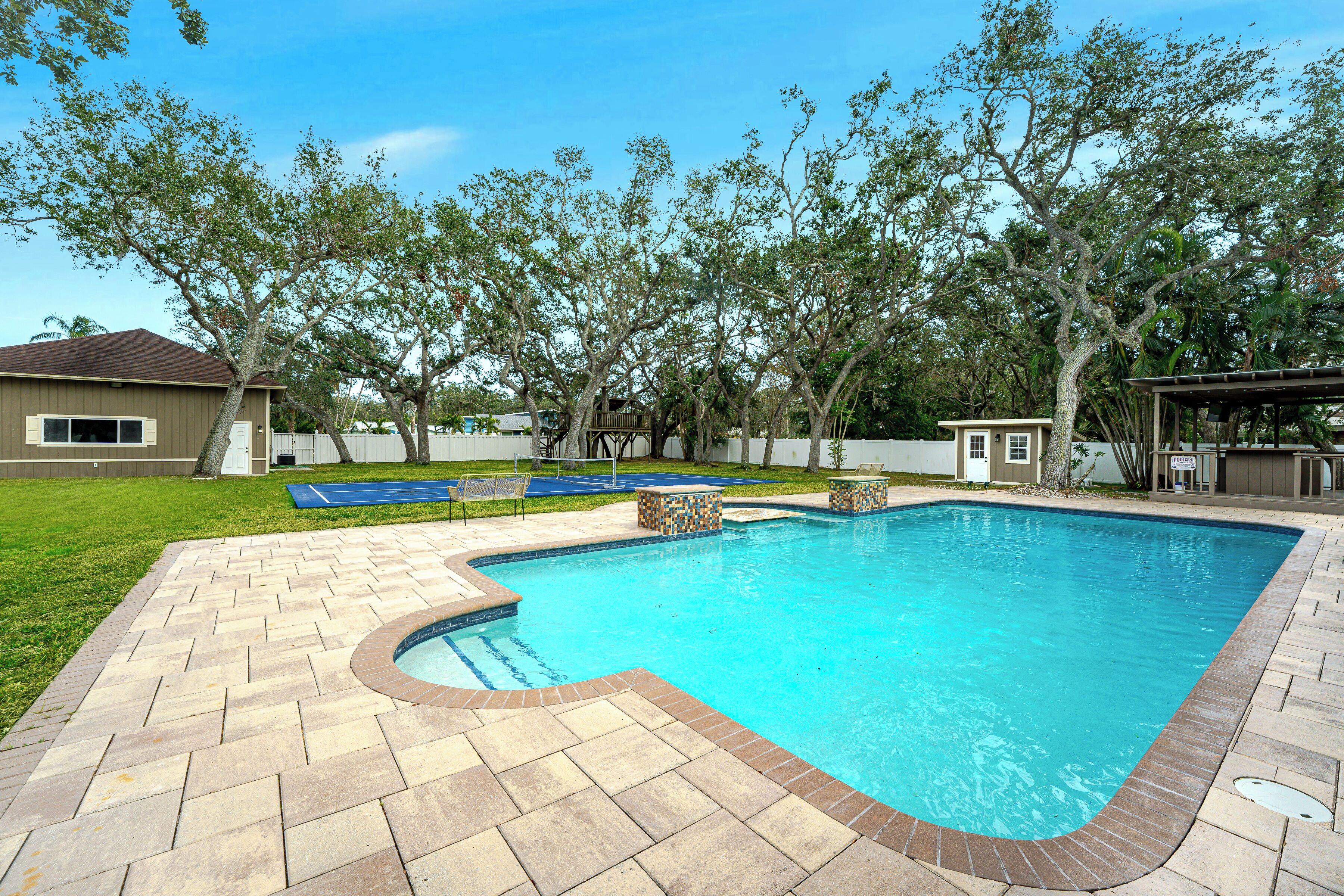 A rectangular swimming pool with a herringbone tile pattern around the edges, surrounded by a grassy lawn and trees.