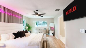 Een moderne slaapkamer met een bed, nachtkastje, plafondventilator en een aan de muur bevestigde televisie waarop het Netflix-logo te zien is.