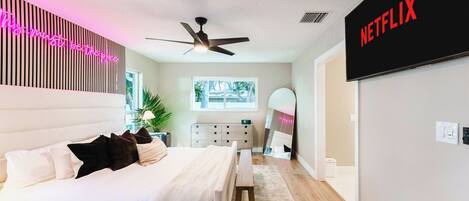 A modern bedroom with a bed, bedside table, ceiling fan, and a wall-mounted TV displaying the Netflix logo.
