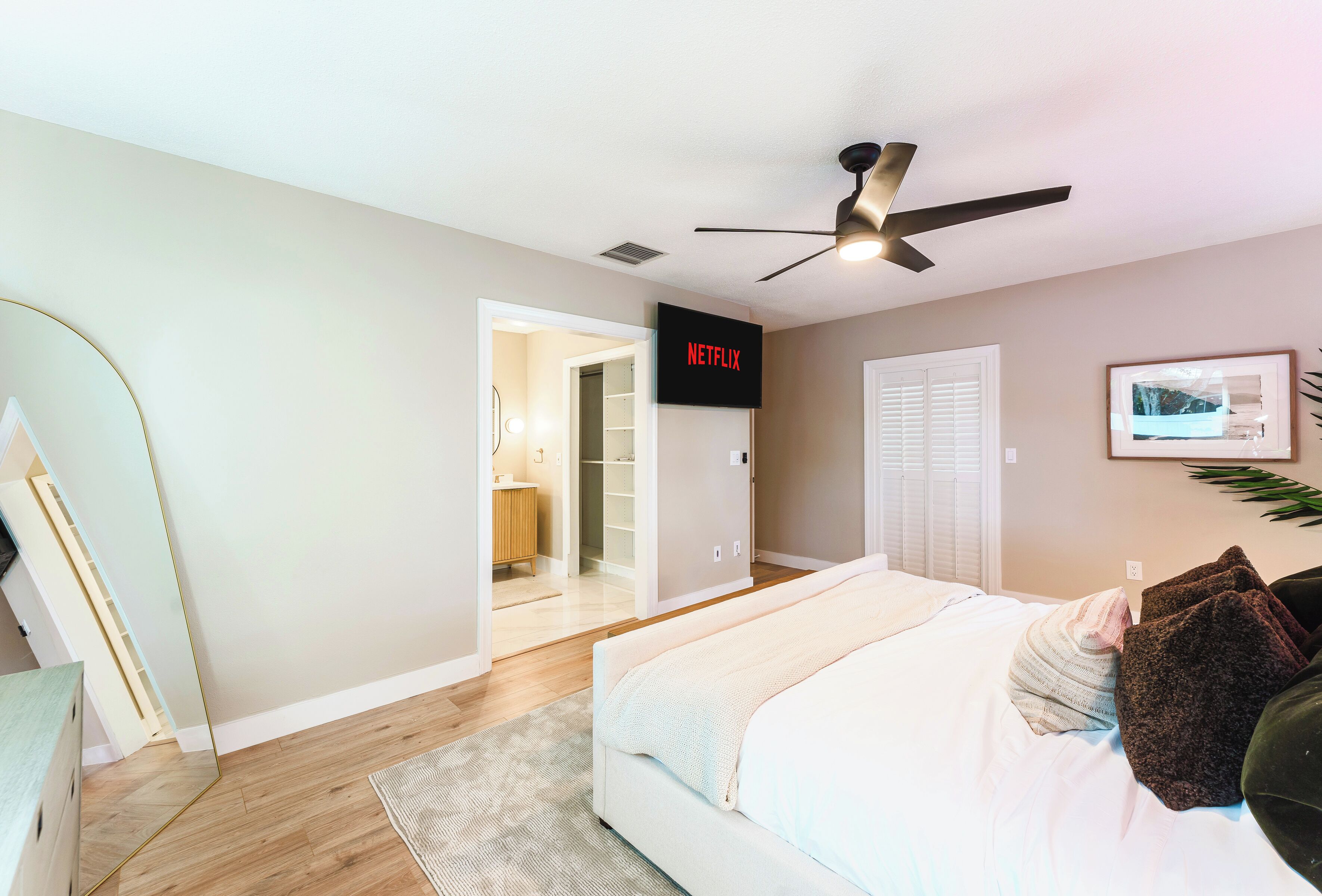 A bedroom with a bed, a ceiling fan, a TV mounted on the wall, and a door leading to a bathroom.