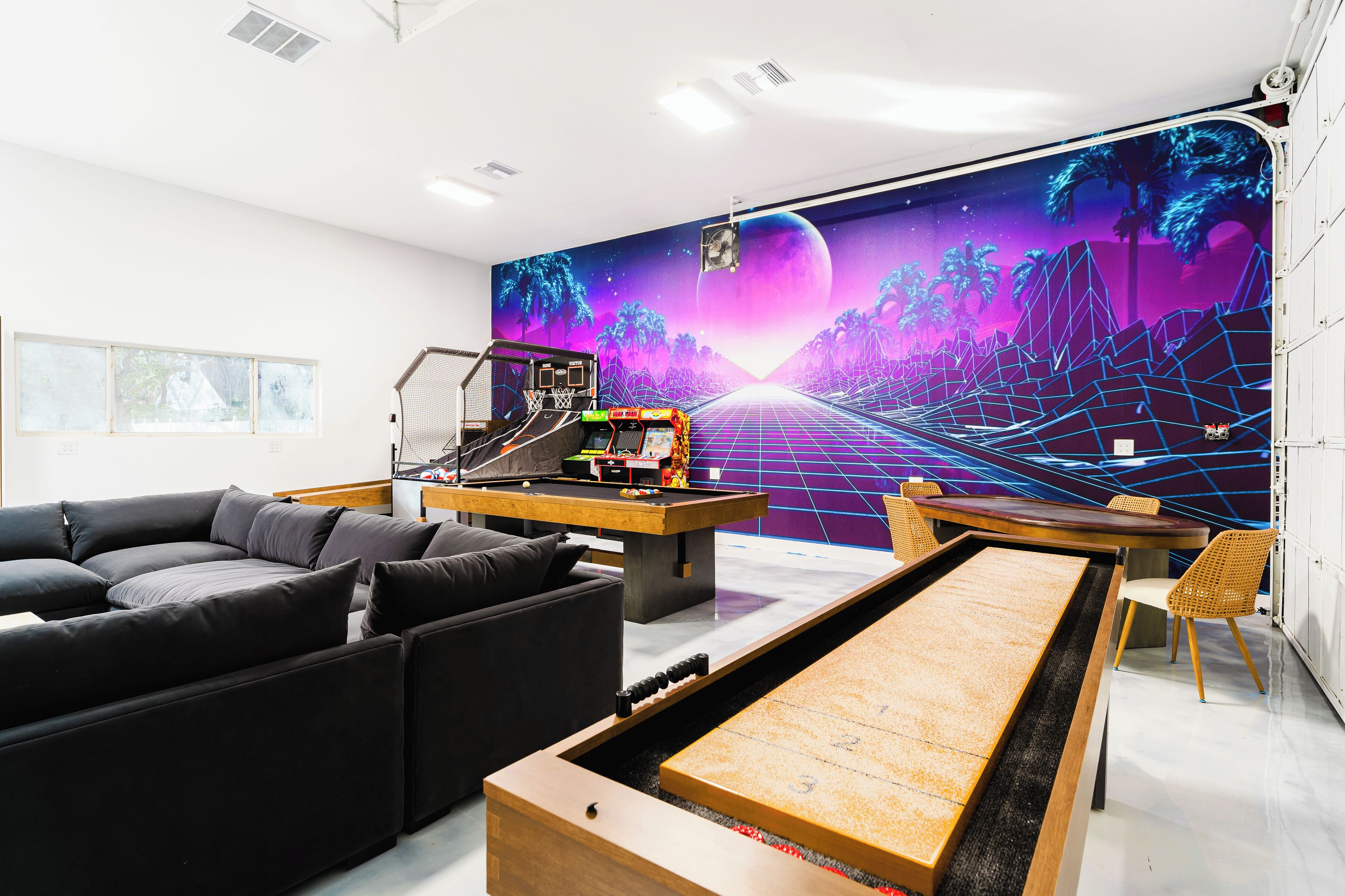 A modern living room with a large sofa, a coffee table, and a gaming table. A mural with a futuristic landscape is on the wall.