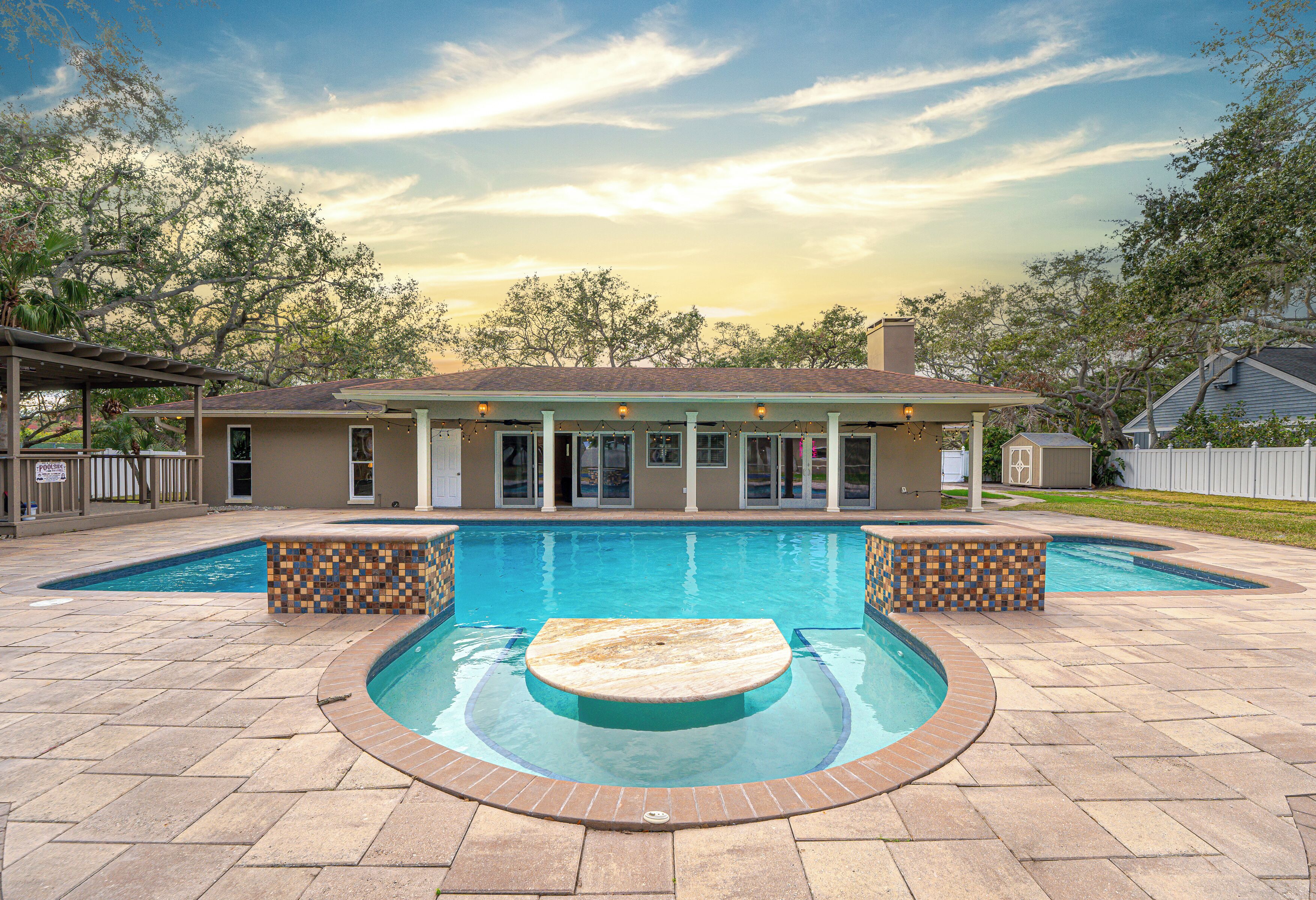 A swimming pool with a sunken center, surrounded by a brick deck, and a house in the background.