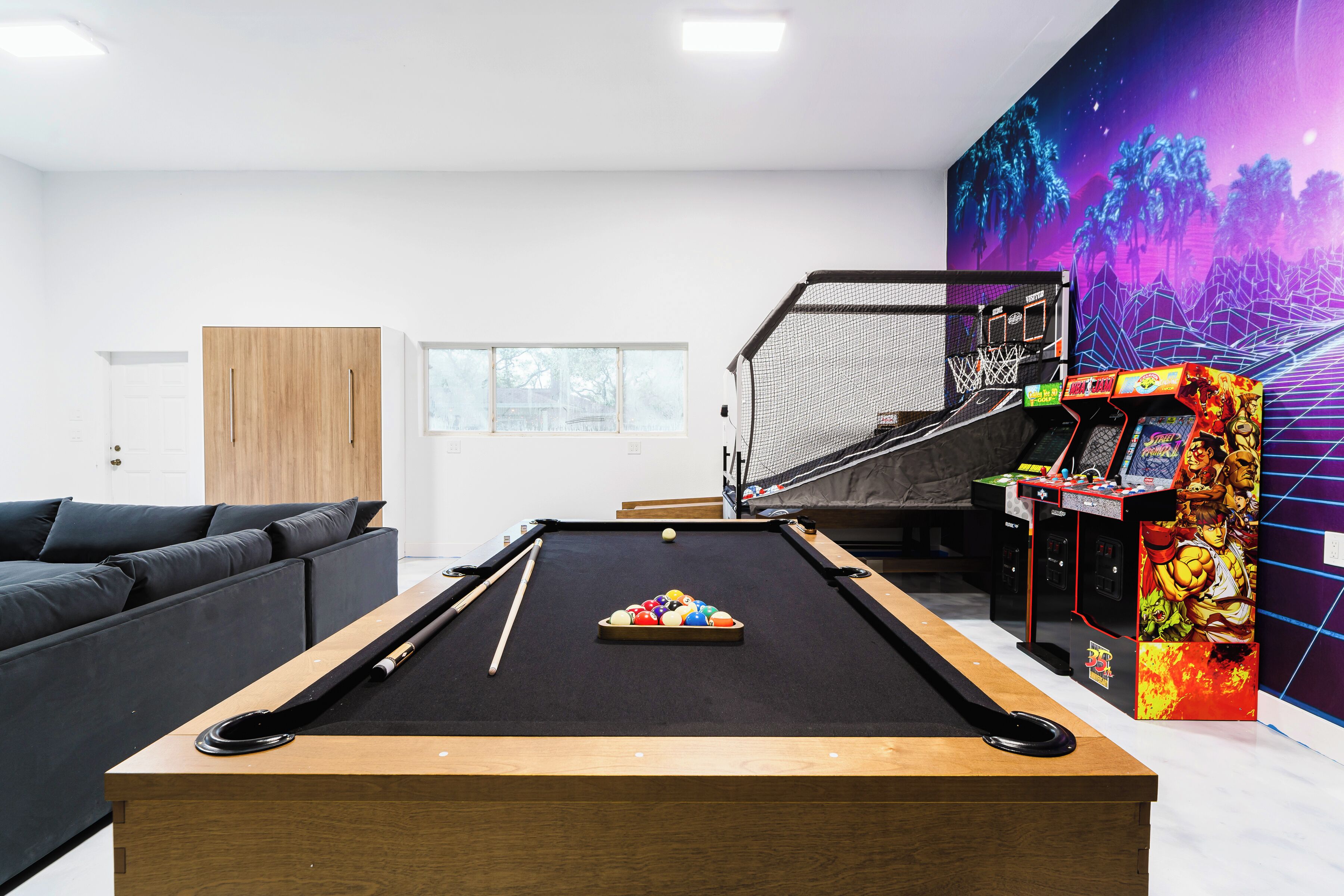 Une table de billard avec un ensemble de billes aux couleurs vives, un canapé, un meuble en bois et une borne d’arcade rétro.