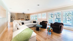 A spacious living room with a large sectional sofa, a small round coffee table, and a colorful ottoman. There are two dark armchairs and a fireplace. The room has large windows with a view of a body of water.