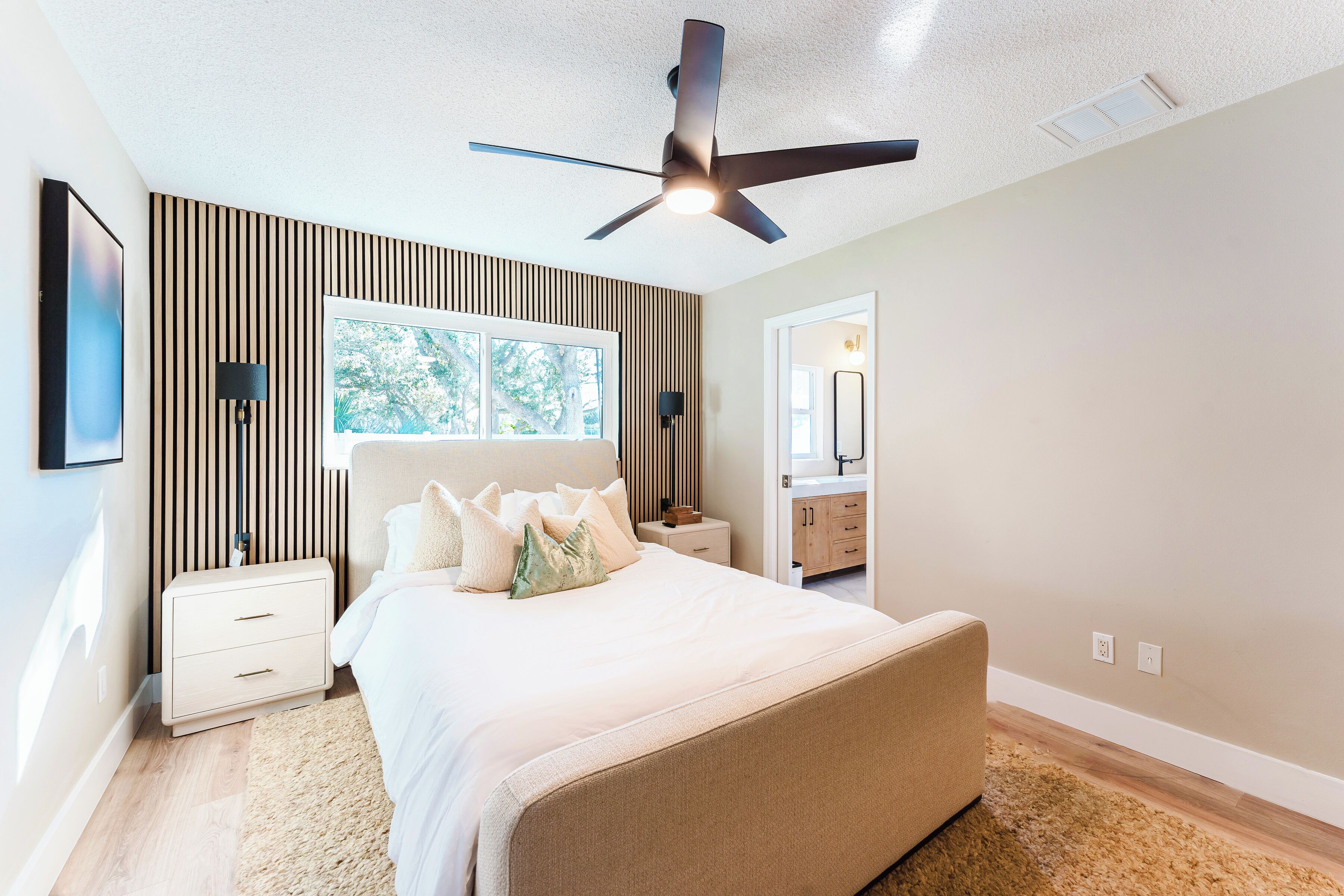 A bedroom with a bed, a ceiling fan, a nightstand, a window, and a bathroom visible through an open door.