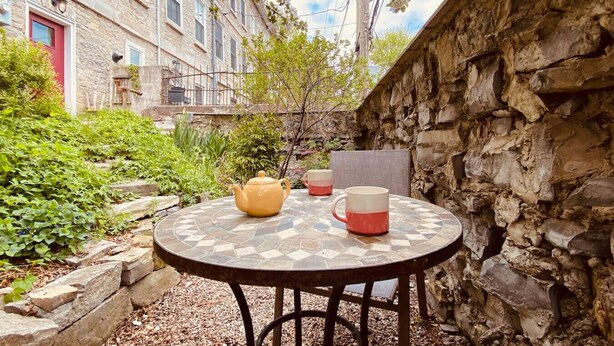 A stone wall, a round table with a teapot and mugs, and a building with windows and a red door in the background.
