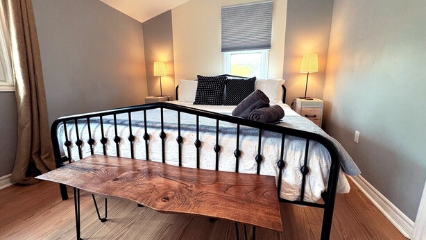 A bedroom with a bed, two bedside lamps, a bench, and a window with blinds. - The Neighbourhood: Expansive loft | free parking (Kingston)