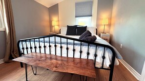 A bedroom with a bed, two bedside lamps, a bench, and a window with blinds.