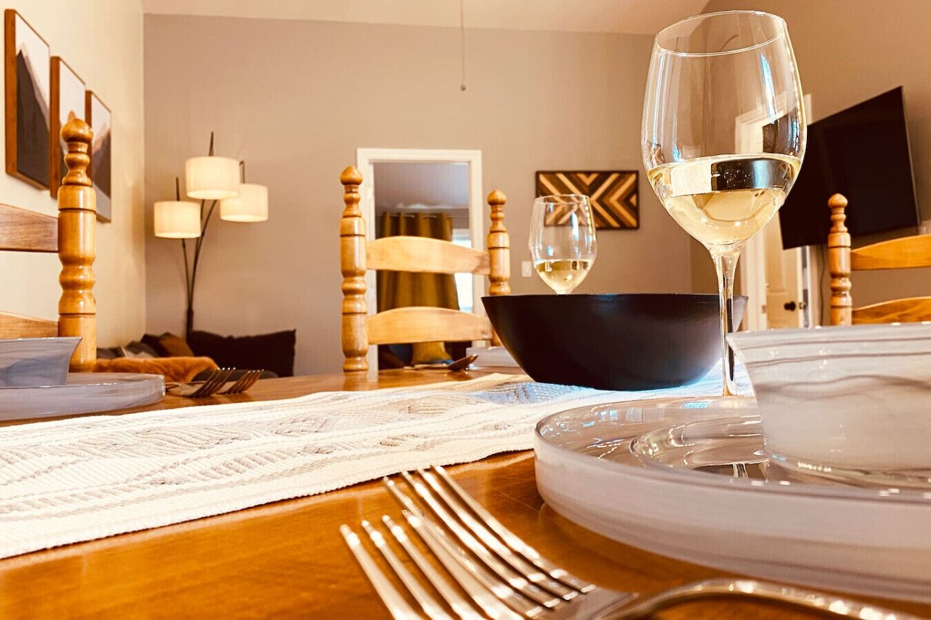 Un comedor con una mesa de madera dispuesta para una comida, un tazón negro, una copa de vino blanco y un plato blanco.