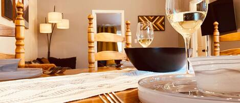 Un comedor con una mesa de madera dispuesta para una comida, un tazón negro, una copa de vino blanco y un plato blanco.