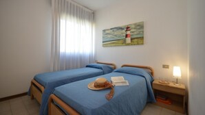 A room with two beds, a painting of a lighthouse, a window with curtains, and a bedside table with a lamp. - Soak Up the Sun Relaxing Apartment Near the Beach (San Michele al Tagliamento)